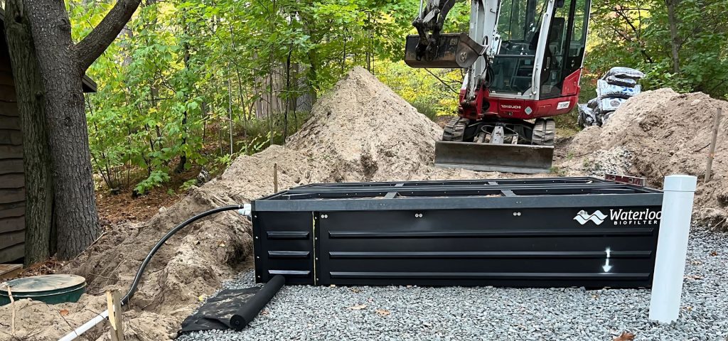 A large black Waterloo Biofilter septic tank is installed in a gravel-covered area. A small red and white excavator sits nearby, with a dirt mound behind it. Trees and green foliage surround the clearing, indicating a wooded location. Greenleaf Excavation Muskoka
