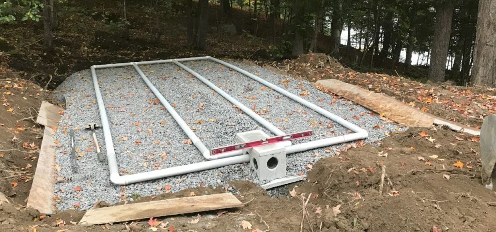A septic drain field under construction in a wooded area; four white PVC pipes are laid horizontally over a bed of gravel, with soil and wooden boards at the edges and a pink level tool resting on a central concrete distribution box. Fallen leaves cover the ground. Greenleaf Excavation Muskoka