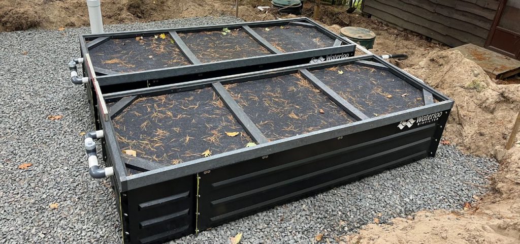 A large, rectangular, black filtration system sits on gravel outdoors, partially surrounded by dirt. The system has two main sections with mesh covers, scattered leaves, and visible piping on the left. Wooden siding and vegetation are in the background. Greenleaf Excavation Muskoka
