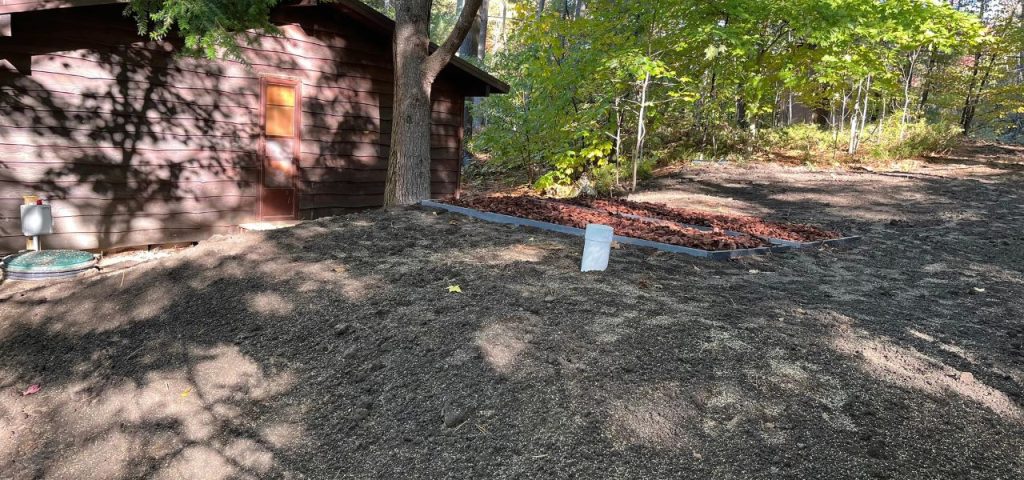 A brown wooden shed stands in dappled sunlight on the left, next to a tree. In front, there’s a patch of bare, seeded soil. To the right, a rectangular garden bed with mulch and a white marker is bordered by gray edging, surrounded by trees with green leaves. Greenleaf Excavation Muskoka