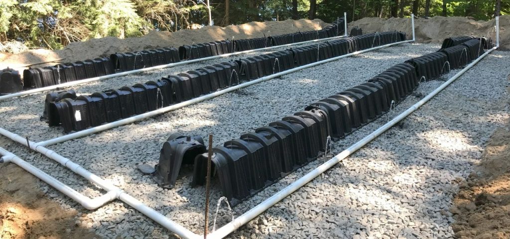 A septic drain field under construction features rows of black plastic chambers laid on gravel beds, surrounded by white PVC pipes. The area is bordered by mounds of soil, with trees and greenery visible in the background. Greenleaf Excavation Muskoka