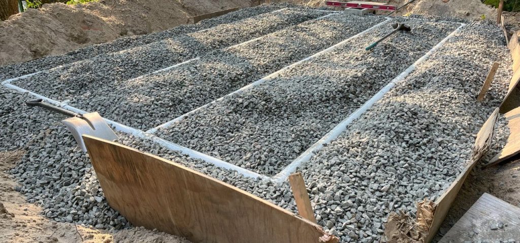 A rectangular construction form filled with gray gravel sits in a dirt area. Wooden boards outline the edges and divide sections. Shovels and leveling tools are placed on top, and piles of dirt surround the form, indicating ongoing construction work. Greenleaf Excavation Muskoka A rectangular construction form filled with gray gravel sits in a dirt area. Wooden boards outline the edges and divide sections. Shovels and leveling tools are placed on top, and piles of dirt surround the form, indicating ongoing construction work. Greenleaf Excavation Muskoka