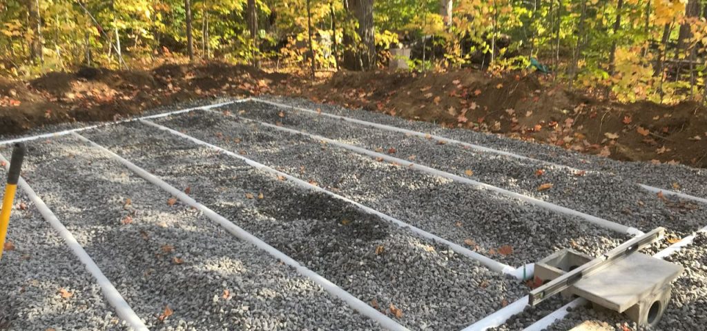 A gravel-covered area is laid out with parallel white PVC pipes, likely part of a septic system drain field installation. In the background, a dirt mound borders a forested area with colorful autumn foliage. A level and shovel are visible at the edge. Greenleaf Excavation Muskoka A gravel-covered area is laid out with parallel white PVC pipes, likely part of a septic system drain field installation. In the background, a dirt mound borders a forested area with colorful autumn foliage. A level and shovel are visible at the edge. Greenleaf Excavation Muskoka