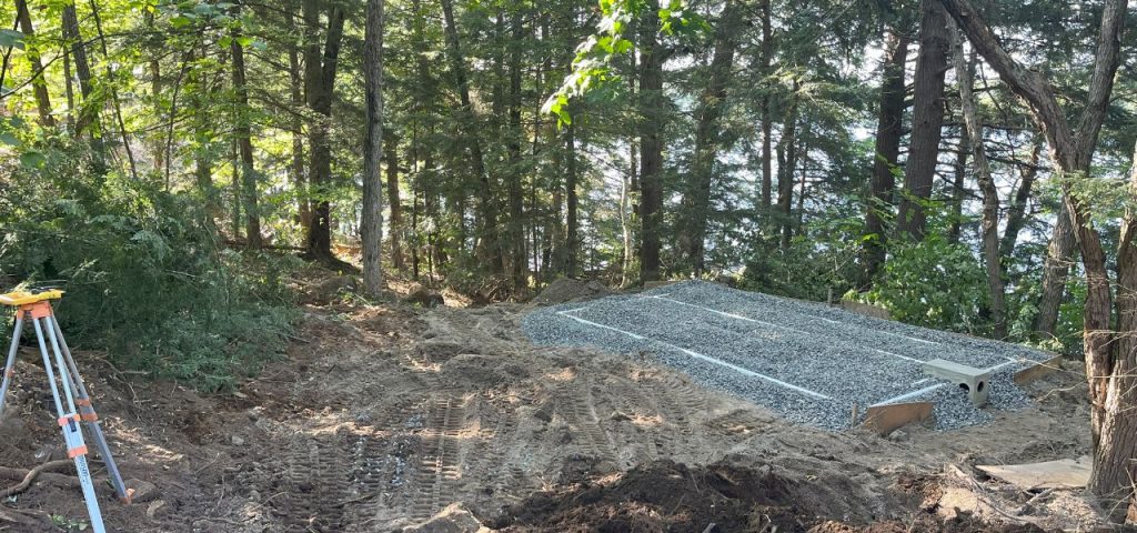 A forested area with sunlight filtering through the trees shows construction work. In the foreground, there are dirt tire tracks and a survey tripod. Midground features a rectangular gravel bed with white pipes, bordered by forest and overlooking a body of water. Greenleaf Excavation Muskoka A forested area with sunlight filtering through the trees shows construction work. In the foreground, there are dirt tire tracks and a survey tripod. Midground features a rectangular gravel bed with white pipes, bordered by forest and overlooking a body of water. Greenleaf Excavation Muskoka