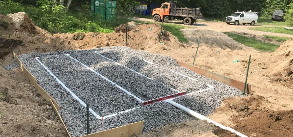 A gravel bed with white PVC pipes arranged in a grid pattern is surrounded by wooden boards and soil piles at a construction site. Trees, a green container, an orange dump truck, a white van, and a black SUV are visible in the background. Greenleaf Excavation Muskoka A gravel bed with white PVC pipes arranged in a grid pattern is surrounded by wooden boards and soil piles at a construction site. Trees, a green container, an orange dump truck, a white van, and a black SUV are visible in the background. Greenleaf Excavation Muskoka