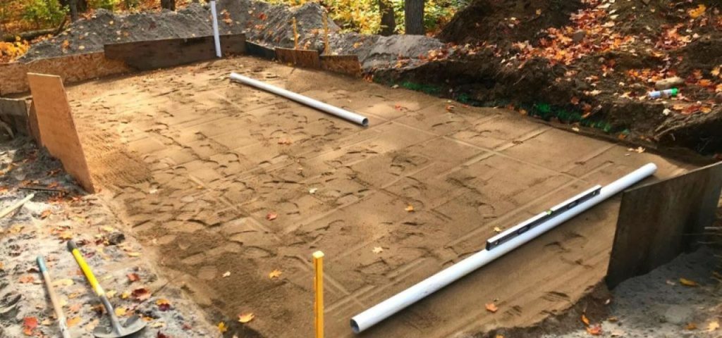 A construction site shows a rectangular, sand-leveled foundation bordered by wooden forms, with two white PVC pipes laid across it. Tools and piles of dirt are nearby, and fallen autumn leaves are scattered around the area. Greenleaf Excavation Muskoka A construction site shows a rectangular, sand-leveled foundation bordered by wooden forms, with two white PVC pipes laid across it. Tools and piles of dirt are nearby, and fallen autumn leaves are scattered around the area. Greenleaf Excavation Muskoka