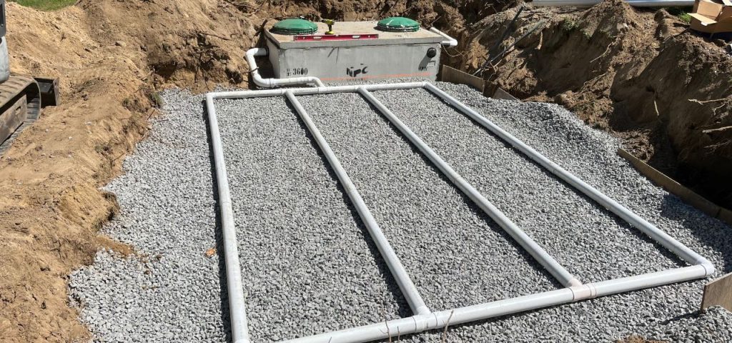 A septic system installation in progress: a rectangular gravel bed with four parallel white PVC pipes is in the foreground, while a concrete septic tank with two green covers and labeled A septic system installation in progress: a rectangular gravel bed with four parallel white PVC pipes is in the foreground, while a concrete septic tank with two green covers and labeled