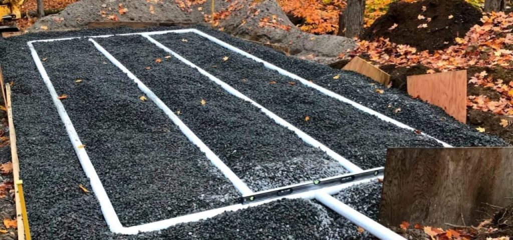 A gravel bed with three white PVC pipes arranged in parallel rows is set outdoors amid fallen autumn leaves. Wooden boards border the area, and soil piles are nearby, indicating a construction site for a drainage or septic system. Greenleaf Excavation Muskoka A gravel bed with three white PVC pipes arranged in parallel rows is set outdoors amid fallen autumn leaves. Wooden boards border the area, and soil piles are nearby, indicating a construction site for a drainage or septic system. Greenleaf Excavation Muskoka