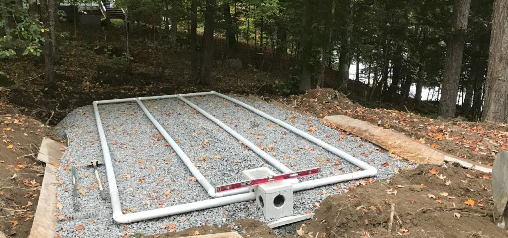 A septic drain field installation in a wooded area, featuring four parallel white PVC pipes laid on a bed of gravel. A spirit level rests on top of the central pipe, and fallen leaves are scattered across the gravel and surrounding soil. Trees and a lake are visible in the background. Greenleaf Excavation Muskoka A septic drain field installation in a wooded area, featuring four parallel white PVC pipes laid on a bed of gravel. A spirit level rests on top of the central pipe, and fallen leaves are scattered across the gravel and surrounding soil. Trees and a lake are visible in the background. Greenleaf Excavation Muskoka
